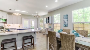 A modern kitchen with white cabinets, a central island with stools, a dining area with wicker chairs, and large windows. - Legacy Villa 2602 - 3 Bedroom 2 Bath With Garage in Beachside Neighborhood (Gulfport)