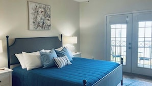 A bedroom with a bed, nightstands, a ceiling fan, a door with glass panels, and a window. - Legacy Villa 203 - 2 Bedroom 2 Bath With Garage in a Beachside Neighborhood (Gulfport)