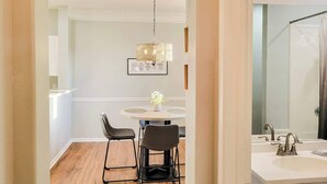 A dining area with a round table, two chairs, a chandelier, and a white wall with a framed picture. - Legacy Villa 1103 - 1 Bedroom 1 Bath With Garage in Beachside Neighborhood (Gulfport)