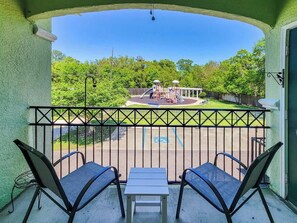A balcony with two chairs and a table, overlooking a playground with a fenced area and trees in the background.