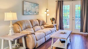 A living room with a beige sofa, two white side tables, a wooden coffee table, a painting on the wall, and a view through a glass door. - Legacy Villa 1103 - 1 Bedroom 1 Bath With Garage in Beachside Neighborhood (Gulfport)