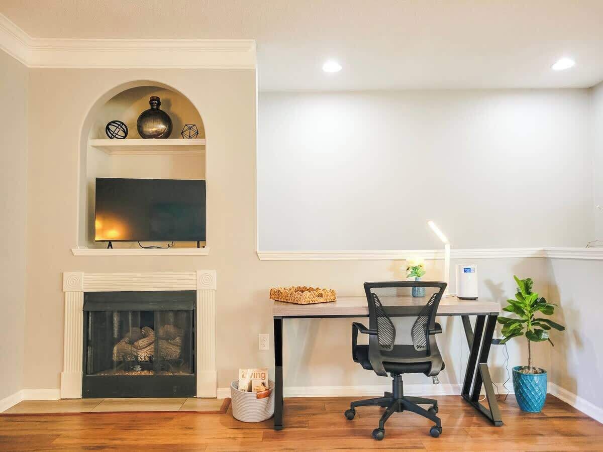 A room with a fireplace, a TV mounted above it, a desk with a chair, and a potted plant.