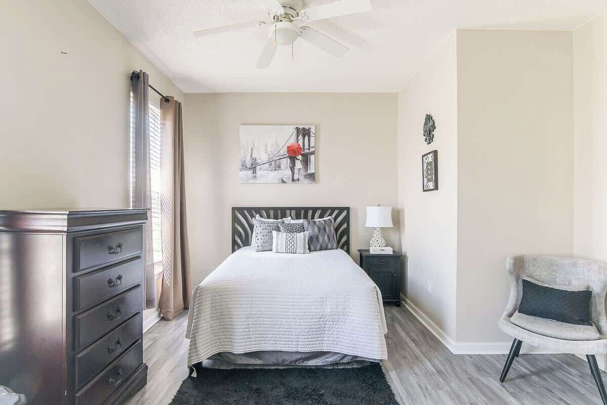 Une chambre à coucher comprenant un lit, une commode, une table de chevet, une chaise et une œuvre d’art accrochée au mur.