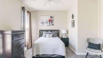Une chambre à coucher comprenant un lit, une commode, une table de chevet, une chaise et une œuvre d’art accrochée au mur.