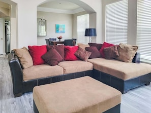 A sectional sofa with multiple cushions, a matching ottoman, a dining table with chairs, and a kitchen area visible in the background.