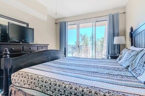 Et hotelværelse med en seng, et fjernsyn på en kommode, en balkon med gardiner og udsigt til palmer.