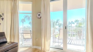 Ein Zimmer mit einer Couch, einer Schiebetür zu einem Balkon mit Gartenmöbeln und Palmen.