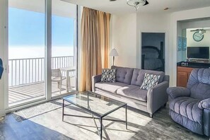 Un salon moderne avec une table basse en verre, un canapé agrémenté de coussins à motifs, un fauteuil inclinable, un téléviseur et un balcon offrant une vue sur l’océan.