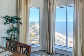 A room with a large window offering a view of a beach and a road. There is a wooden chair and a table with a patterned table runner.