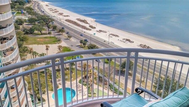 A balcony with a view of a beach and ocean, a pool, and a multi-story building. - The Cobalt View (Gulfport)