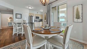 A dining area with a round wooden table, white upholstered chairs, and a chandelier. - The Cobalt View (Gulfport)
