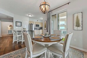 A dining area with a round wooden table, white upholstered chairs, and a chandelier. - Spacious Beach View, End Unit in Legacy Tower 2-1301 - 3 Bedroom 3 Bath Condo (Gulfport)