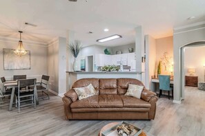 A living room with a brown leather sofa, a dining table with chairs, a chandelier, and a kitchen area in the background.