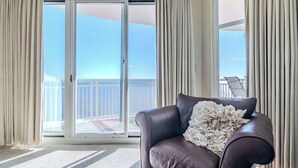Een kamer met een donkerbruine leren bank, een witte geplooide plaid en uitzicht op een balkon met een hekwerk.