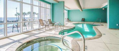 Ett poolområde med två bubbelpooler, en sittgrupp med stolar och stora fönster som erbjuder utsikt över stranden.