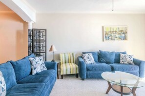 A living room with two blue sofas, a striped chair, a glass coffee table, a lamp, and a painting on the wall. - Beachview Condo in Legacy Tower 1 -1104 - Fully Furnished 2 Bedroom 2 Bath (Gulfport)