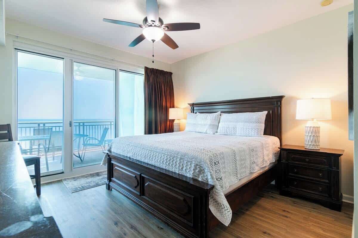 Ein Schlafzimmer mit einem großen Bett, einem Deckenventilator, einem Nachttisch und Blick auf den Balkon.