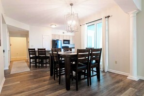 A dining area with a wooden table and chairs, a kitchen in the background, and a large window with curtains. - Spacious Beachview End Unit in Legacy Tower 2 -1101 - 3 Bedroom 3 Bath Condo (Gulfport)