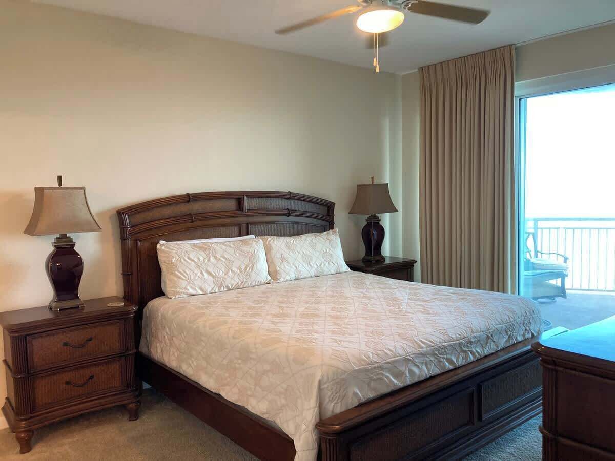 A bedroom with a large bed, two bedside tables with lamps, a ceiling fan, and a view of a balcony.