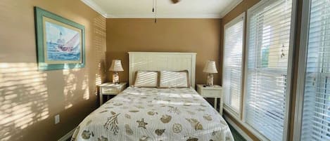 Un dormitorio con cama, ventilador de techo, dos mesitas de noche con lámparas, un cuadro en la pared y ventanas grandes con persianas.