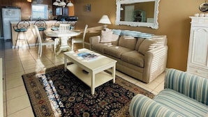 Ein Wohnzimmer mit einem beigefarbenen Sofa, einem gestreiften Sessel, einem Couchtisch und einem gemusterten Teppich. Im Hintergrund ist eine Küche mit Kühlschrank sowie ein Essbereich mit Stühlen zu sehen.