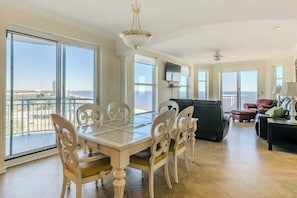 A spacious living area with a dining table, chairs, a sofa, and a TV. Large windows offer a view of the city and a balcony.