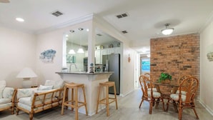A modern kitchen with white cabinets, a central island with stools, and a dining area with wicker chairs. - Cypress Cove 103 - Beautiful 1 Bedroom 1 Bath Beachfront Condo in Biloxi (Biloxi)