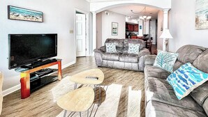A living room with a flat-screen TV, two coffee tables, a grey sofa with patterned cushions, and a rug. - Beachview Condo at Legacy Tower 2 -1104 - Fully Furnished 2 Bedroom 2 Bath (Gulfport)