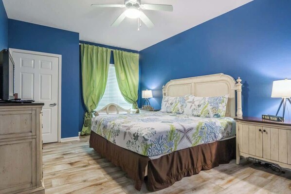 Une chambre à coucher comprenant un lit, une table de chevet, une commode, un ventilateur de plafond et une fenêtre avec des rideaux.