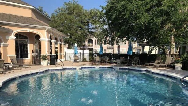 A swimming pool with a 3-foot depth marker, surrounded by lounge chairs and umbrellas, with a building featuring arches in the background. - Legacy Villa 1306 - 2 Bedroom 2 Bath With Garage in Beachside Neighborhood (Gulfport)