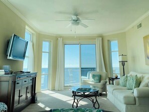 A spacious living room with a television, a ceiling fan, a large window with curtains, a white sofa, and a coffee table. - Spacious Beachview End Unit in Legacy Tower 1 -1001 - 3 Bedroom 3 Bath Condo (Gulfport)