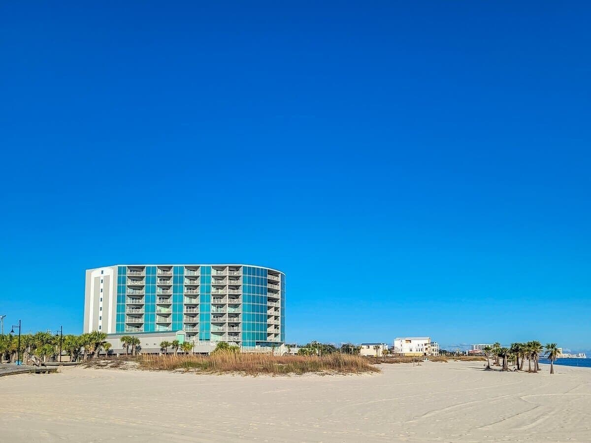 A large, multi-story hotel with a modern design, located near a beach with palm trees and a clear blue sky.