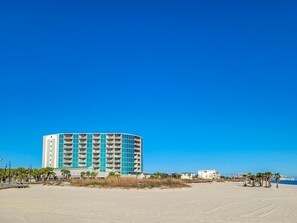 A large, multi-story hotel with a curved facade, located on a sandy beach with palm trees and a clear blue sky.