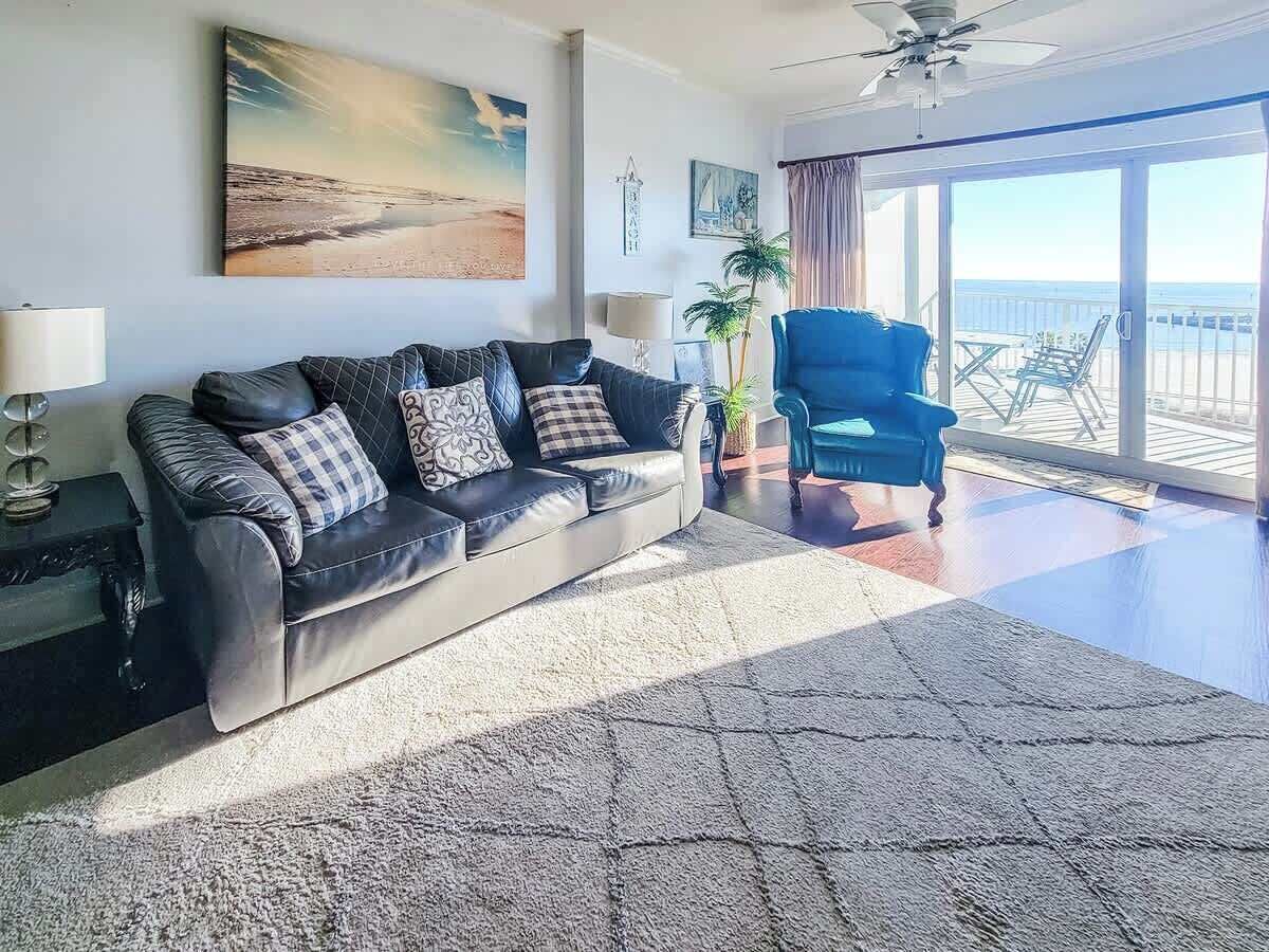 A living room with a sofa, a blue armchair, a large rug, and a view of the ocean through a balcony.
