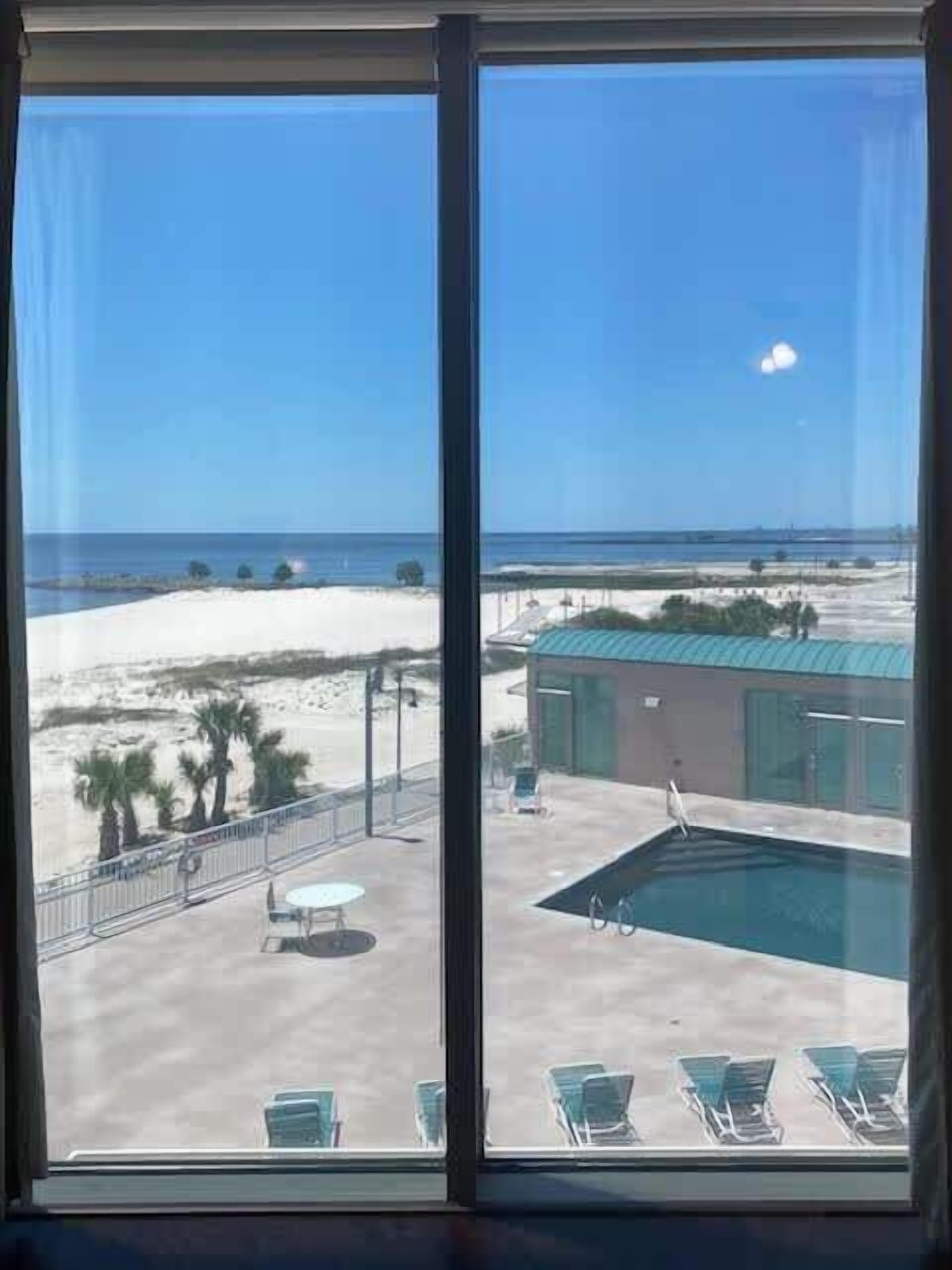 A view from a window overlooking a pool area with a beach and a building in the background.