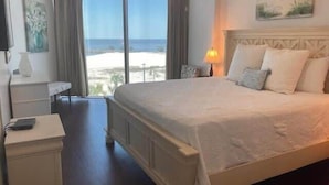 Ett hotellrum med en stor säng, ett nattduksbord, en lampa och utsikt över stranden.