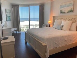 A hotel room with a large bed, a bedside table, a lamp, and a view of the beach.
