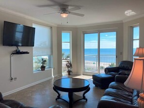 A spacious living room with a large TV, a ceiling fan, and a view of the ocean through large windows. - Spacious Beachview End Unit in Legacy Tower 1-501 - 3 Bedroom 3 Bath Condo (Gulfport)