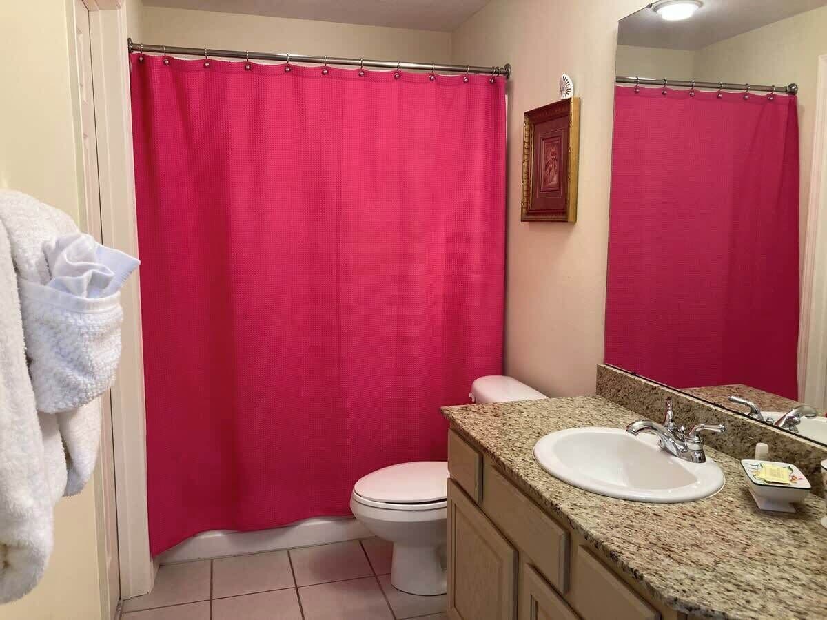 A bathroom with a pink shower curtain, a white toilet, a granite countertop sink, and a mirror.