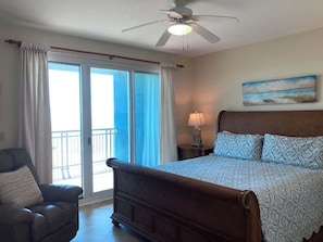 A bedroom with a large bed, a chair, a nightstand with a lamp, a ceiling fan, and a painting on the wall.