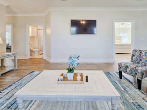 A living room with a large flat-screen TV mounted on the wall, a coffee table with a plant, and a patterned armchair. - Brand New Home With Beach Views! Fully Furnished 3 Bedroom Home in Gulfport (Gulfport)
