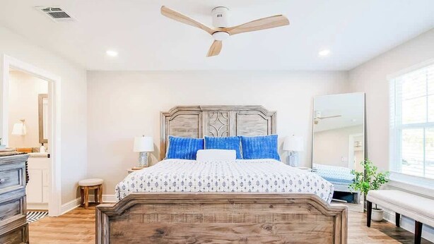A bedroom with a wooden headboard, a bed with blue and white bedding, a ceiling fan, a bedside table with a lamp, and a large window. - Brand New, Fully Furnished 3 Bedroom, 3 Bathroom Home in Ocean Springs! (Ocean Springs)