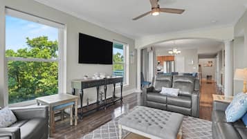 A spacious living room with a large flat-screen TV, a ceiling fan, and a view of the outdoors.