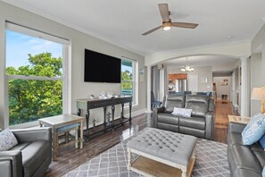 Un salón espacioso con una gran televisión de pantalla plana, un ventilador de techo y vistas al exterior.