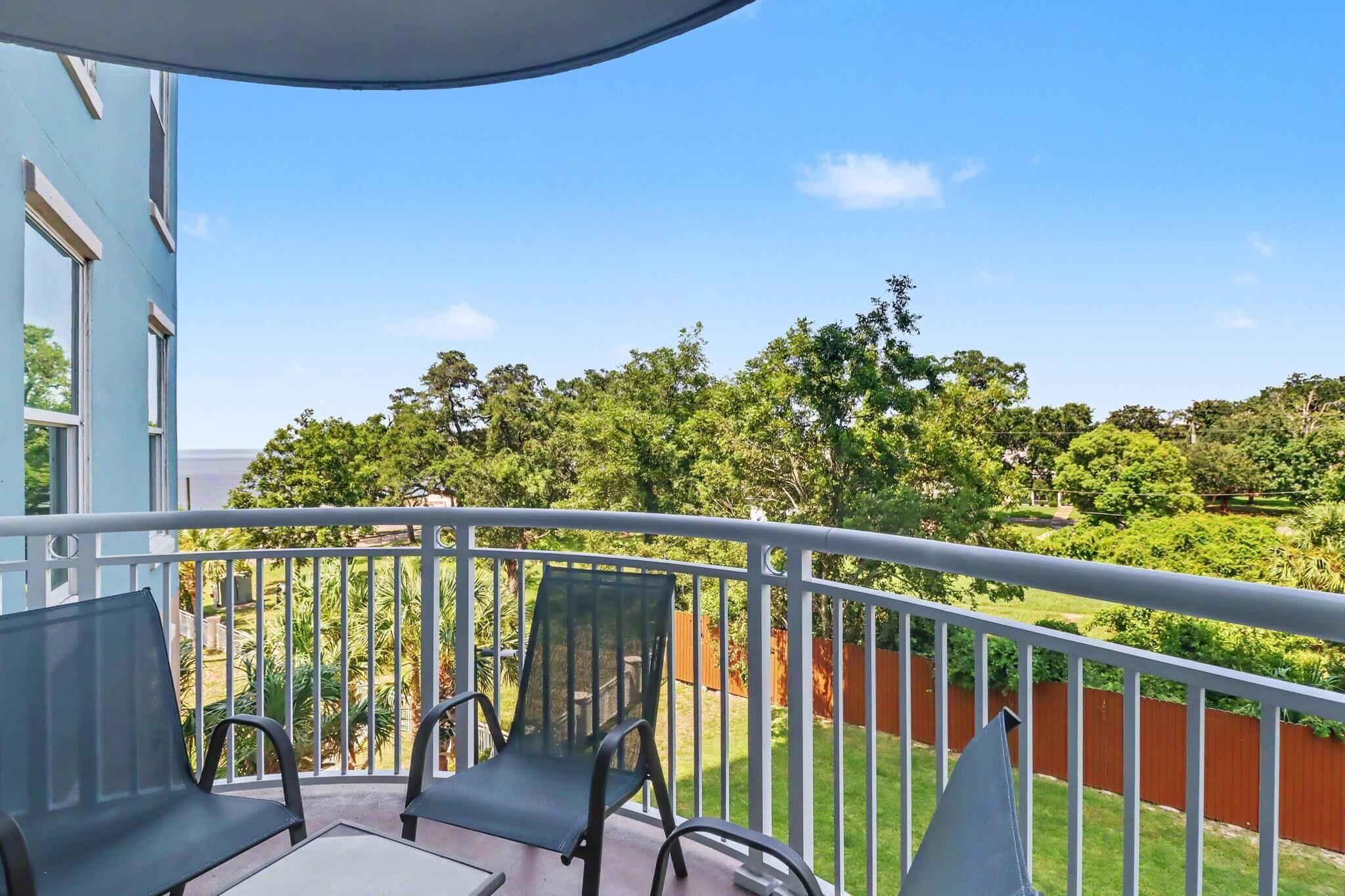 A balcony with a table and chairs, overlooking a garden with trees and a fence.