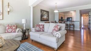 A living room with a sofa and a coffee table, a kitchen in the background, and a hallway leading to another room.