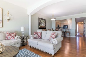 A living room with a sofa, a coffee table, and a rug. There is a kitchen in the background with wooden cabinets and a microwave.