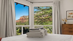 Un dormitorio con un ventanal grande, una cama con ropa de cama blanca, un aparador de madera y un cuadro en la pared.