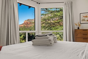 A bedroom with a large window, a bed with white linens, a wooden dresser, and a framed picture on the wall.