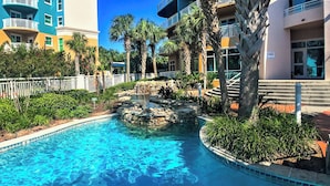 A swimming pool with a rock feature surrounded by palm trees and a fence. - Beachview Condo at Legacy Tower 2 -1103 - Fully Furnished 2 Bedroom 2 Bath (Gulfport)